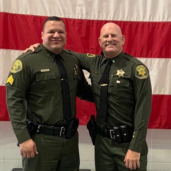 Two Sheriff Deputies in uniform smiling with arms around each other in front of a large American flag.