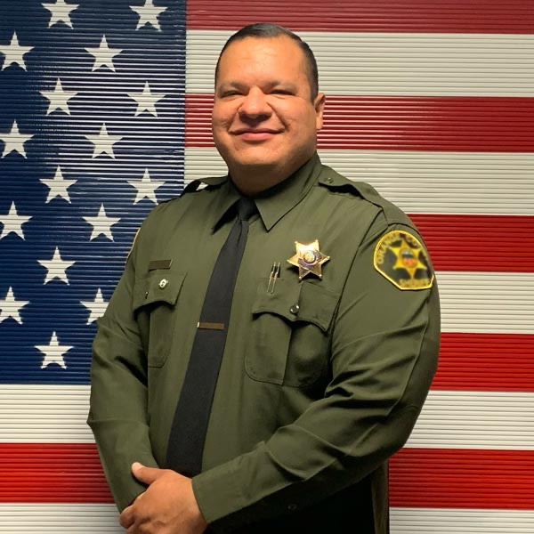 Professional portrait of a smiling Sheriff Deputy (Mike) in uniform, standing in front of an American flag.